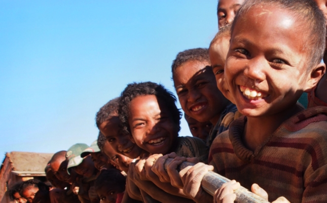 Smiling children hold onto a water pipe to be laid, with bright blue sky behind them.